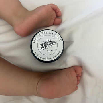 Baby balm container with 'Sol Shade Skin Co.' branding next to a baby's feet on white fabric.