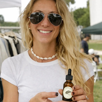 Woman holding a brown bottle with a label, wearing sunglasses and a white shirt outdoors.
