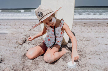 Child playing on a sandy beach with a surfboard in the background