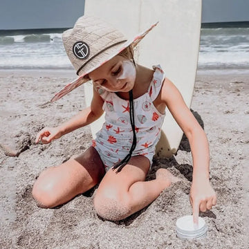 Child sitting on a sandy beach with a surfboard, wearing a hat and playing with sand.